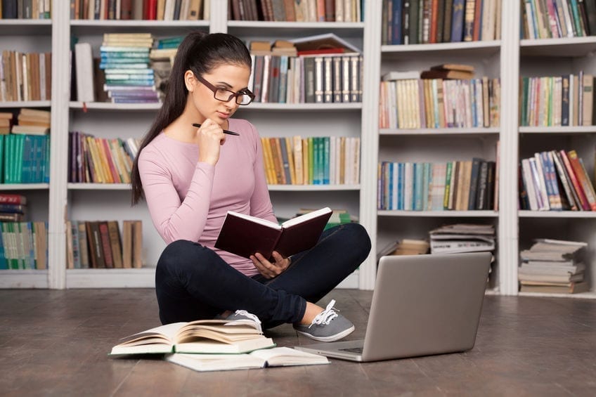 Preparing to the exams. Thoughtful young woman holding a book and looking at the laptop while sitting on the floor at the library Preparing to the exams. Thoughtful young woman holding a book and looking at the laptop while sitting on the floor at the library
